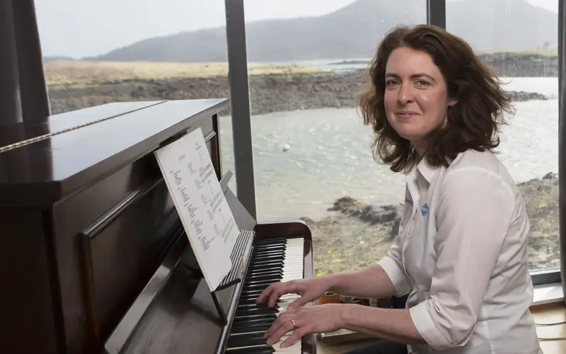Music teacher Olwen Macleod sitting at a piano with island view background