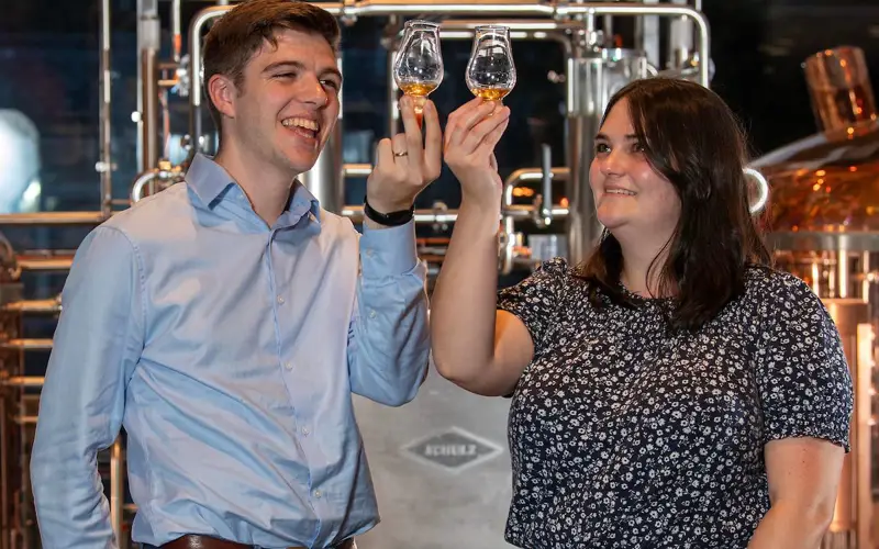 Couple standing in front of a chrome and brass still holding up small glasses of whisky to the light
